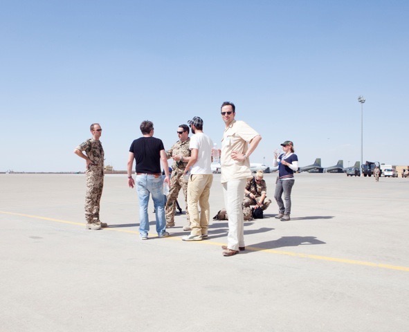 Sometimes the passion for music is reflected in a simple plain airport. A group of people stand loosely around with sunglasses, some of them wearing uniforms, others plainly dressed. Planes can be seen in the background.