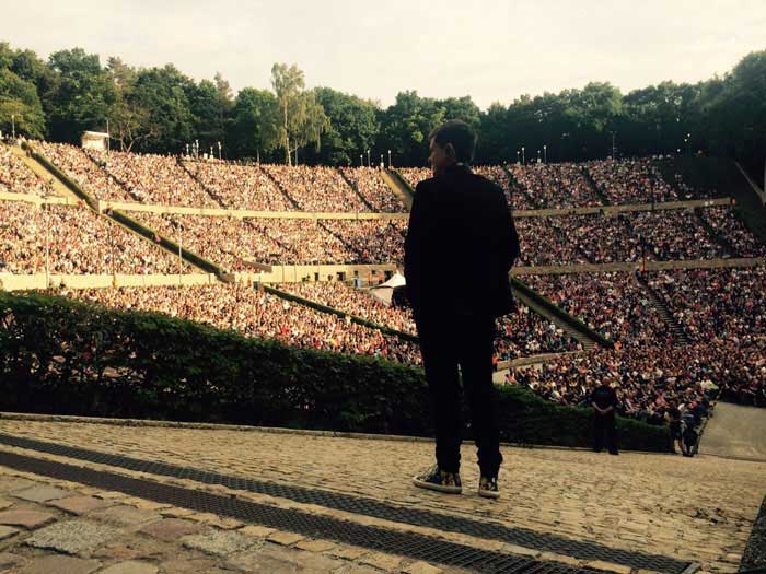 A person stands casually with his hands in his trouser pockets. The person is at the highest point of a steep stony path. Her gaze is fixed on the crowded tiers of the amphitheatre in front of her. She is wearing a mixture of suit and sports shoes.