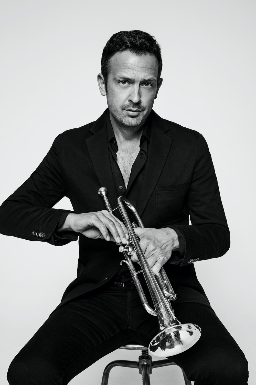 The musician Till Brönner is sitting on a stool. He is fixing something on his trumpet. His shirt is casually open and he looks directly into the camera.